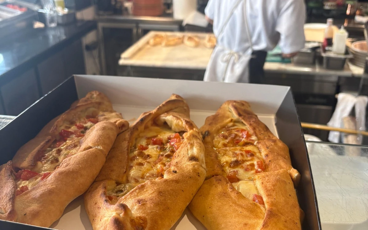 Three boat-shaped Greek pizzas with melted cheese and tomato in a box, a cook working behind