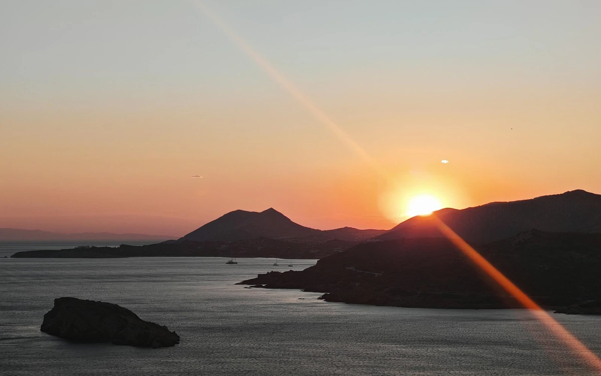 Die tief stehende Sonne über dunklen Hügeln jenseits des Wassers vor Cape Sounion, im Vordergrund eine kleine Felsinsel