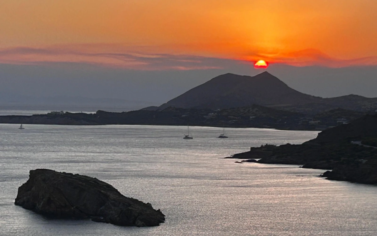 Ein orangefarbener Himmel über dem Saronic Gulf, die Sonne berührt einen Hügelkamm, Segelboote liegen in der Bucht vor Anker