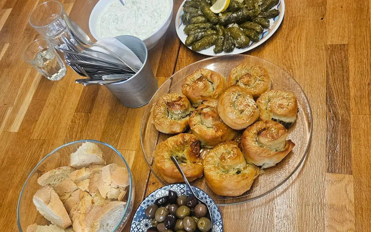A table of Greek dishes: tzatziki, stuffed vine leaves, rolled filo pies, bread and olives